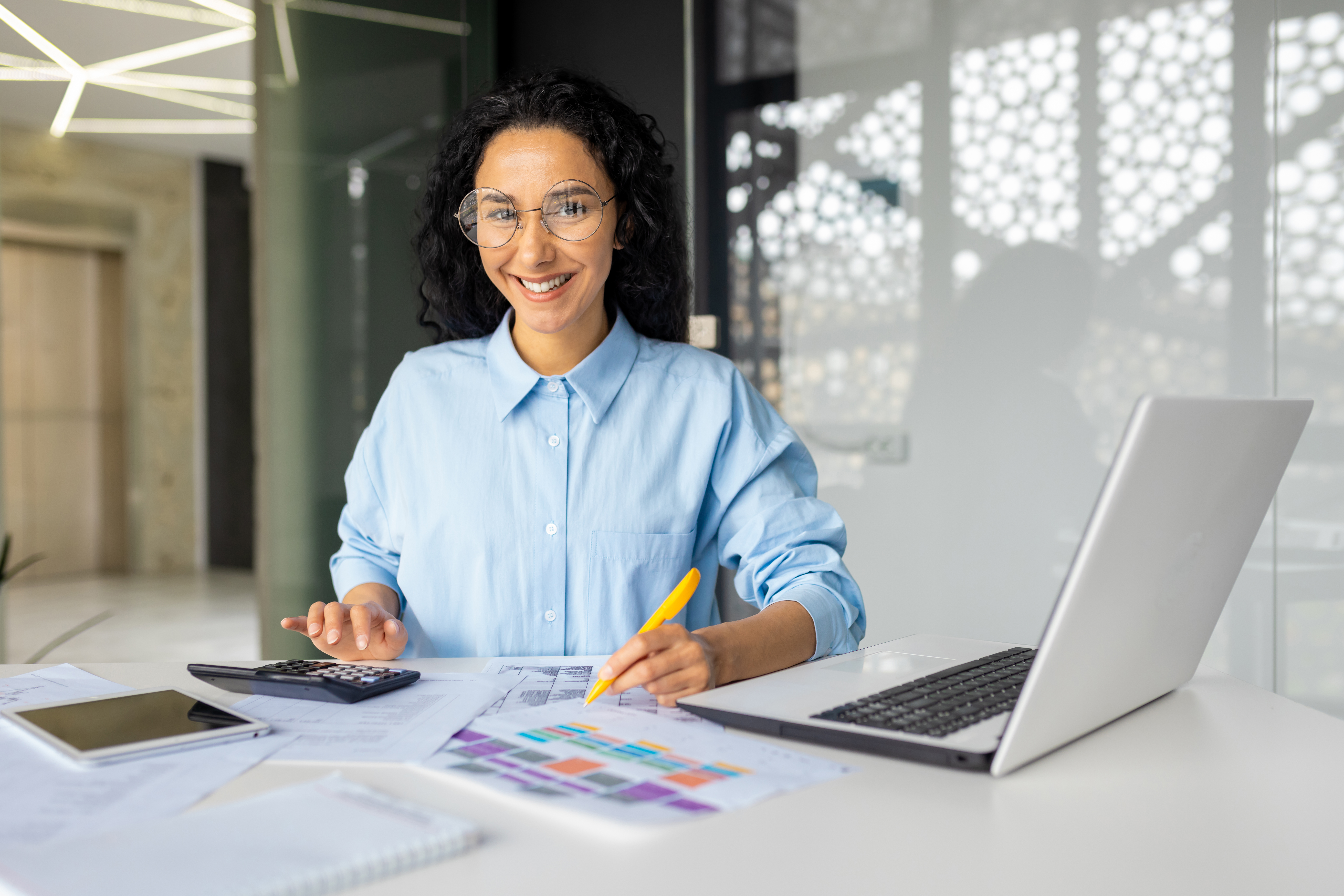 Portrait of young successful financier at workplace inside office, businesswoman fills tables reports and contracts, female worker on paper work with laptop looking at camera smiling